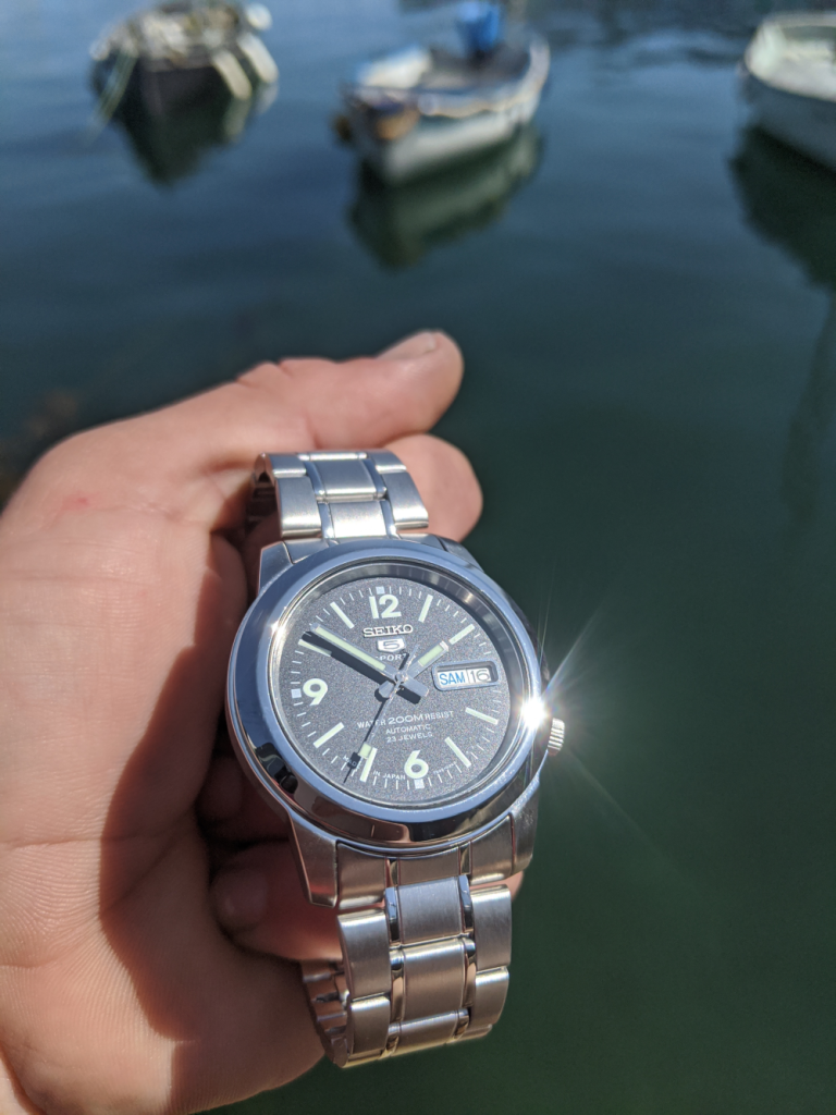 Seiko SNKK Mod - Displayed in clenched palm of hand against a back drop of small skiffs moored in the harbour. Custom Seiko. 