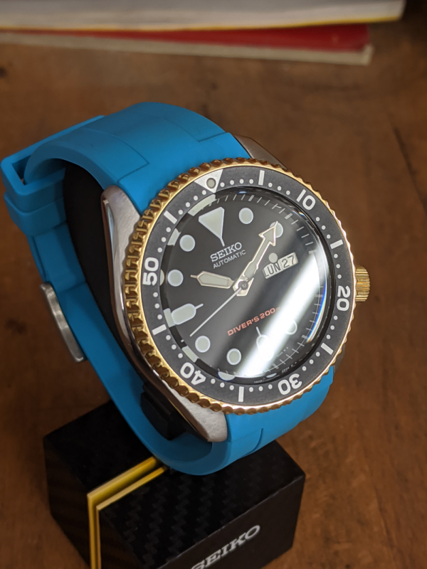 Seiko SKX Mod Gold Plated bezel and crown completed with a light blue strap displayed ona carbon seiko display stand ontop of a wooden work bench