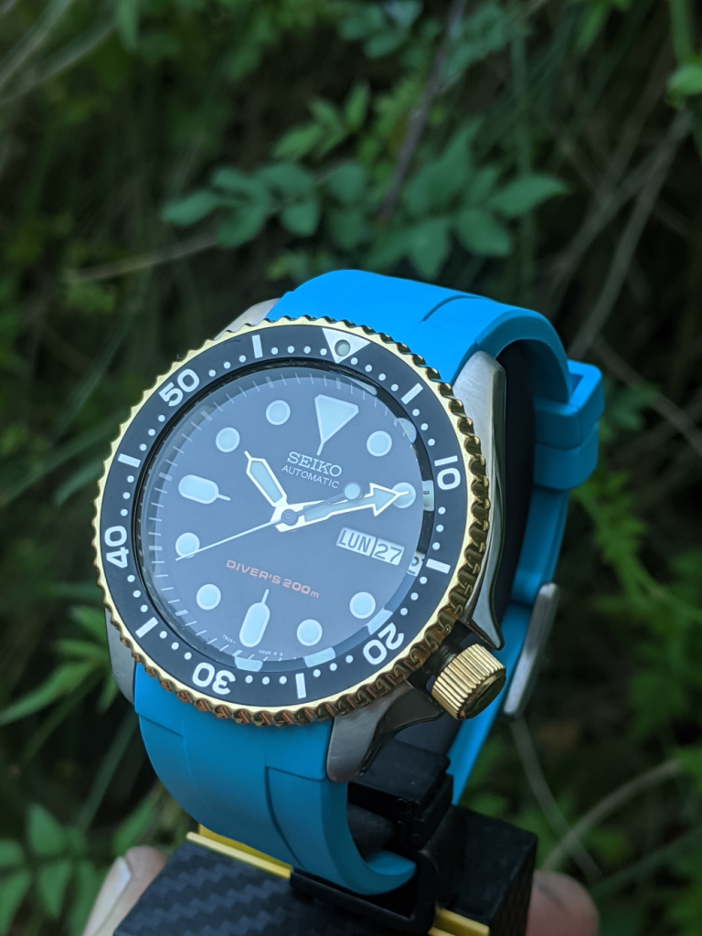 OEM Seiko SKX with 18K Gold Plated bezel and crown, completed with a 2 peice electric blue rubber strap. Displayed amoung the bushes and shrubs on a carbon seiko display stand