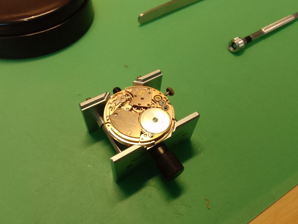 Vintage watch repair - omega Cal 1030 with it is displayed in a bergeon movement holder on on a watchmakers anti-static matt,with a bergeon screwdriver & wach cushion and dupont tweezers