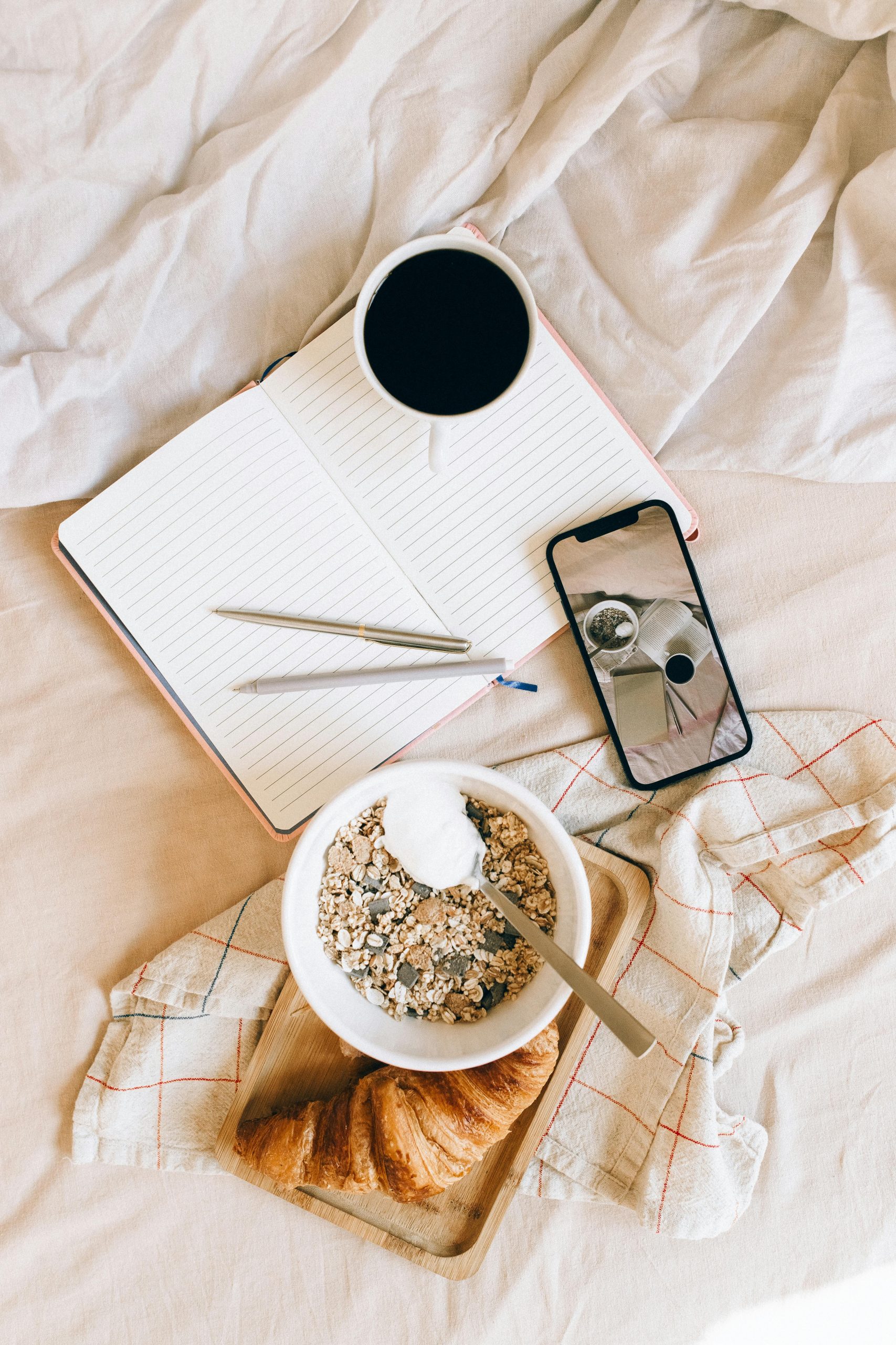 Top view of cozy breakfast setup with coffee, notebook, and smartphone on a bed.