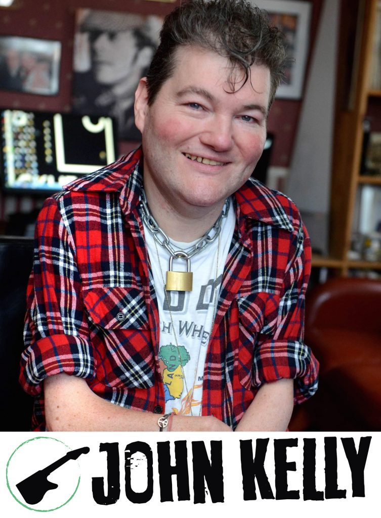 Image Description: John Kelly, a white man wearing a red checked shirt over the top of a vintage t-shirt, sits smiling at the camera. You can see his music technology in the background. John is also wearing a fabulous necklace, which is a heavy chain with a real bronze padlock on it. John is very cool! Underneath John is a white banner which shows the name 'JOHN KELLY' alongside a silhouette of his custom-made musical instrument, called 'The Kellycaster'.