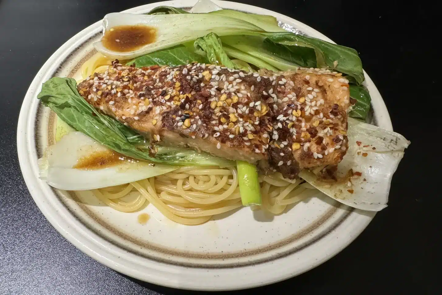 Bed of green leaved vegetables on spaghetti topped with cooked salmon with sprinkled sesame seeds on top