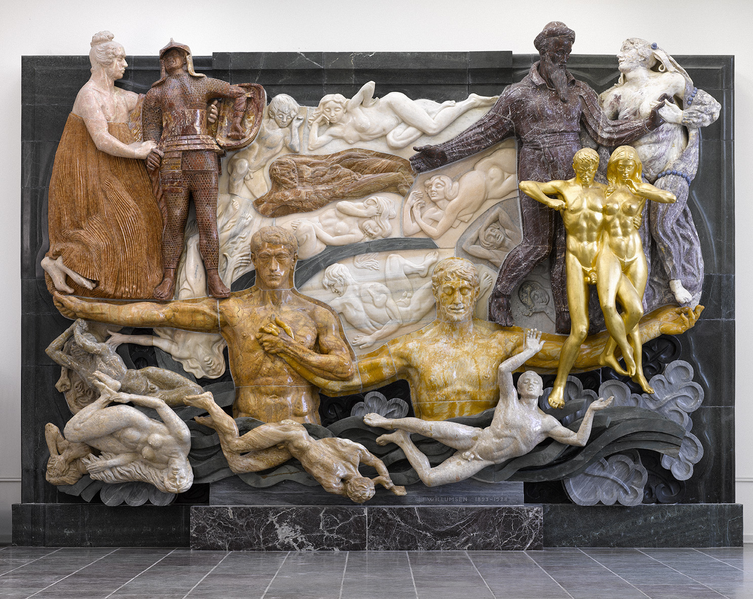 Et stort, indviklet skulptur med flere menneskefigurer i forskellige positurer. Skulpturen har en blanding af materialer og farver, herunder guld, gyldne farver og hvid. Der er to gyldne figurer på højre side, flere figurer i gyldne farver i midten og venstre side, og hvide figurer nederst. Baggrunden inkluderer detaljerede udskæringer af yderligere menneskelige former i relief. Hele stykket er sat mod en mørk stenbaggrund med et flisegulv foran.