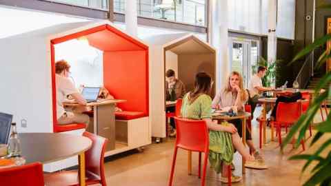 Persons in an indoor office space conversing and/or working on something on their laptops