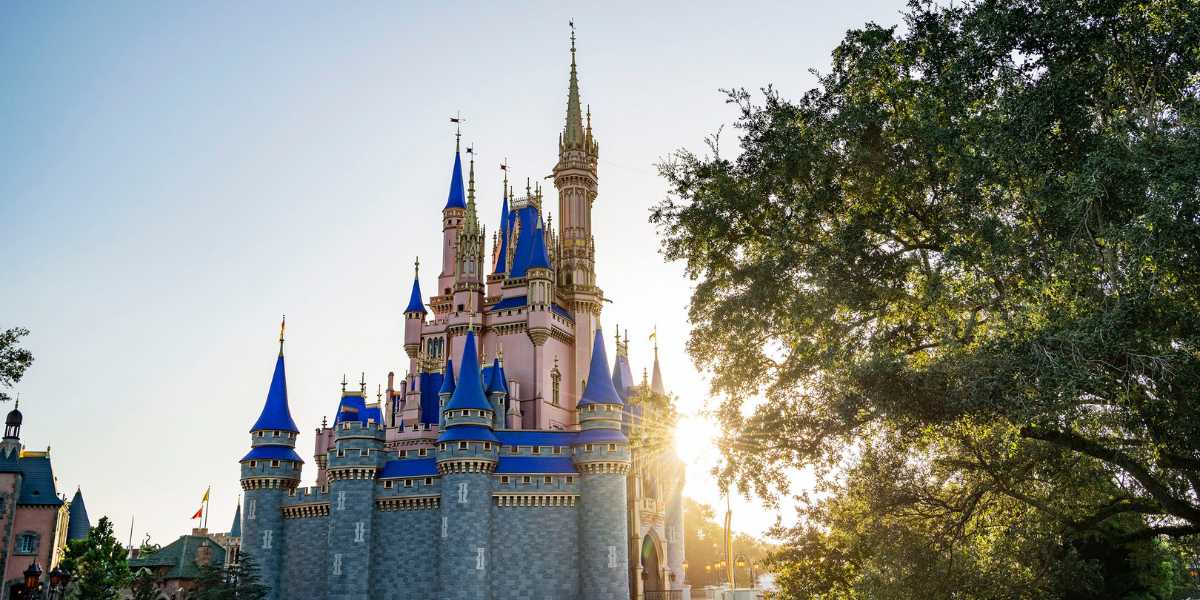 A picturesque view of a fairy tale Cinderella Castle with spires, under a clear blue sky with the sun setting behind, casting a warm glow over the scene.