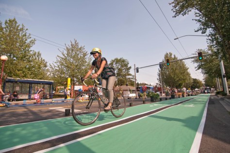 A person wearing a helmet and sunglasses rides a bicycle in a dedicated green bike lane at an intersection under a traffic light showing green. The scene is lively, with people in the background enjoying the street, which is closed to cars and lined with temporary barriers, plants, and decorations.