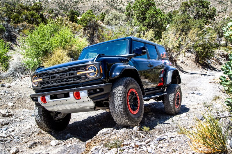 2024 Ford Bronco Raptor Code Orange descending a rocky grade with desert hills and vegetation.