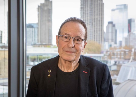 Peter sitting inside, in front of a cityscape - he is wearing a black blazer and t-shirt and black rimmed glasses