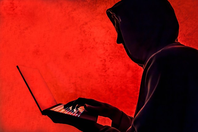 A man wearing a hooded jacket and typing on a laptop, against an eerie red background