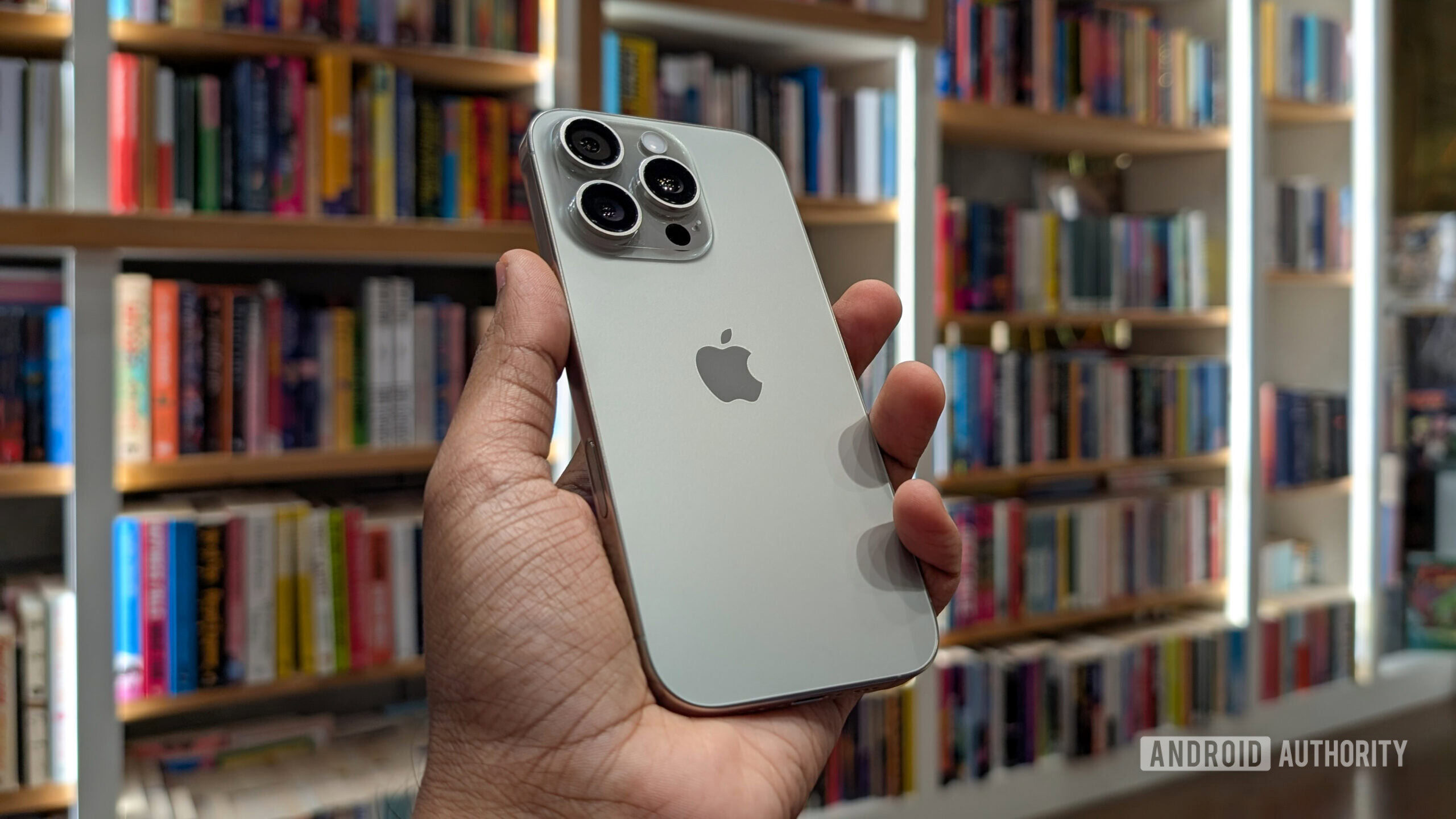 Apple iPhone 16 Pro in Natural Titanium color held out in hand, against the backdrop of books