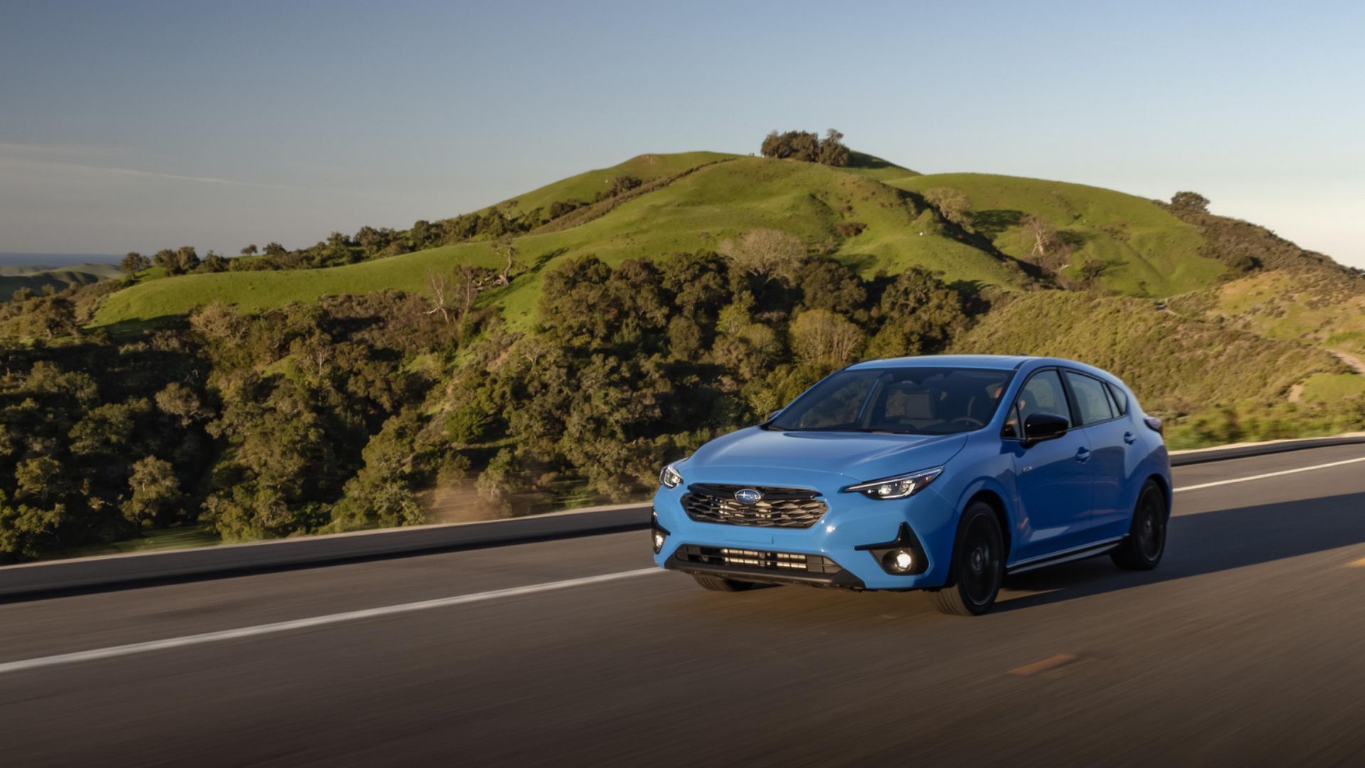 2024 Subaru Impreza RS in blue posing on coastal road with grassy hill in background