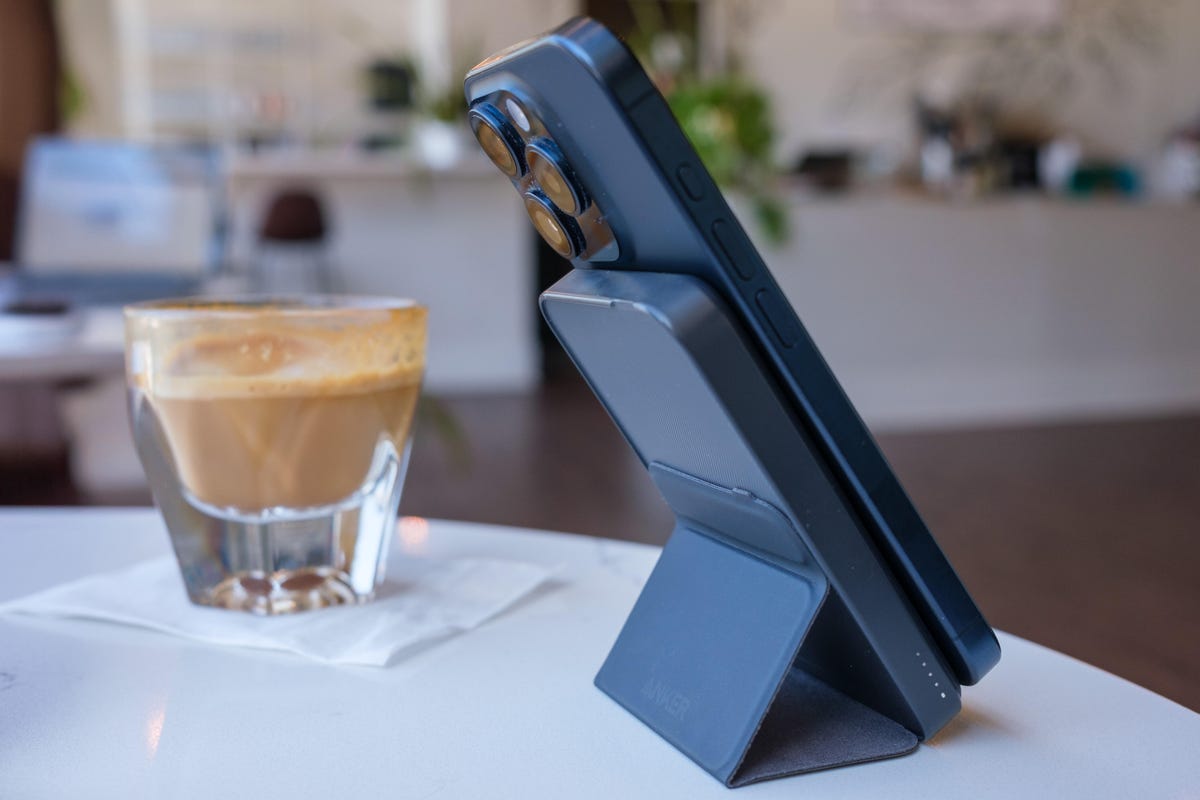 iPhone connected to a slim portable charger on a stand. They're both on a table in a brightly lit cafe along with a cortado in a glass.