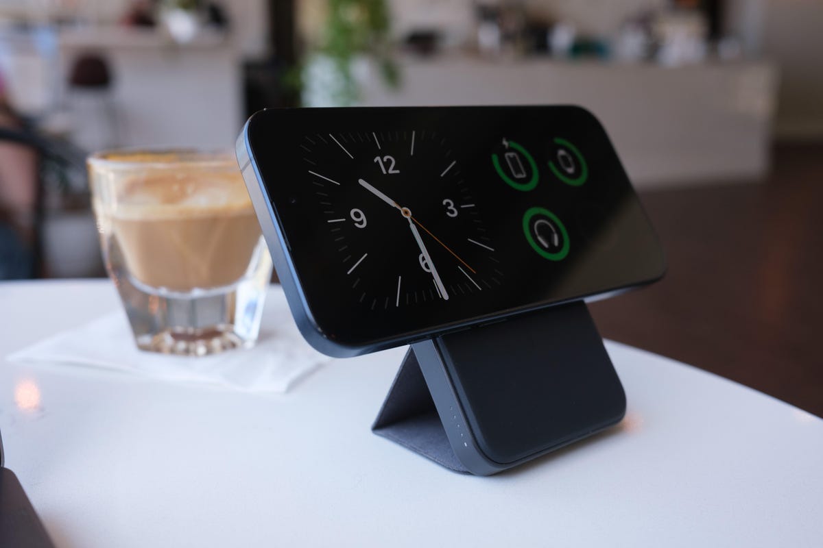 iPhone connected to a slim portable charger on a stand. The phone is oriented horizontally to activate the StandBy mode that shows the current time and other information. They're both on a table in a brightly lit cafe along with a cortado in a glass.