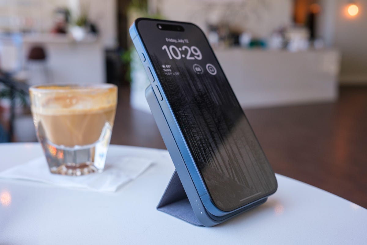 iPhone connected to a slim portable charger on a stand. They're both on a table in a brightly lit cafe along with a cortado in a glass.