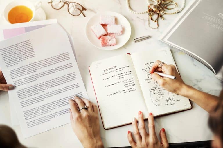 Person writing notes in a notebook with papers and sweets on desk.