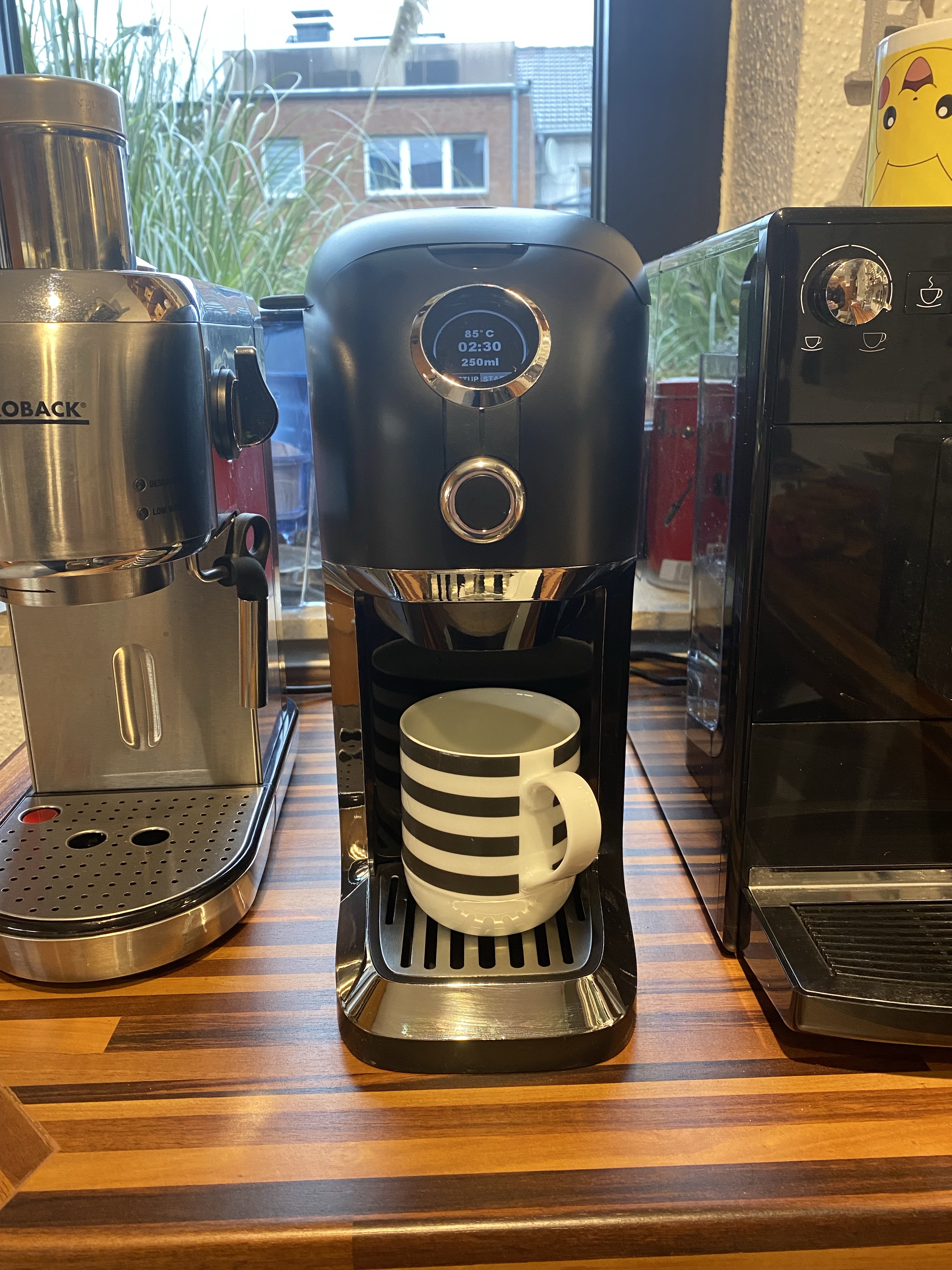Die schwarz-chromfarbene BRU Teemaschine mit schwarz-weiß gestreifter 250ml Tasse zwischen einem Gastrobak Siebträger und einem Melitta Kaffeevollautomaten.