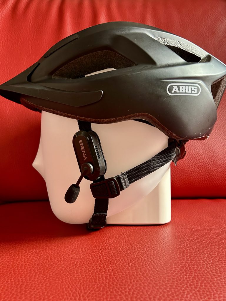 **Alt-Text:** Ein weißer, gesichtsloser Mannequin-Kopf trägt einen schwarzen Fahrradhelm. Auf der rechten Seite des Helms ist ein Mikrofon befestigt, das als Sena BiKom 20 gekennzeichnet ist. Der Hintergrund besteht aus rotem, strukturiertem Kunstleder.