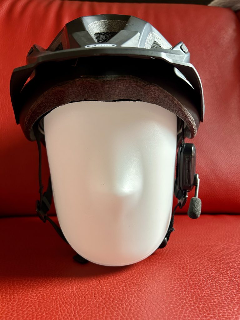 Weißer, gesichtsloser Mannequinkopf mit einem modernen schwarzen Fahrradhelm, der über eine integrierte Freisprecheinrichtung und ein an der rechten Seite befestigtes Mikrofon, das Sena BiKom 20, verfügt. Im Hintergrund ist eine rote, strukturierte Oberfläche zu erkennen, die an ein Möbelstück, etwa ein Sofa oder einen Stuhl, erinnert.