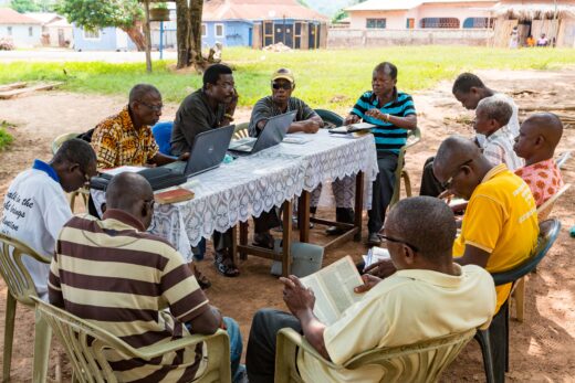 Translators David Livingstone Amoadzah (left, at table, orange/brown shirt) and Togbe Afare VIII (middle, at table, black shirt) sit with reviewers from the Tafi community and discuss the clarity of the New Testament translation work that has been done so far.