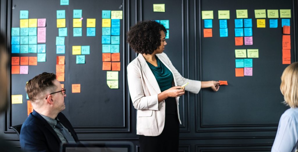 A person standing in front of a whiteboard moving a post it note in a team meeting