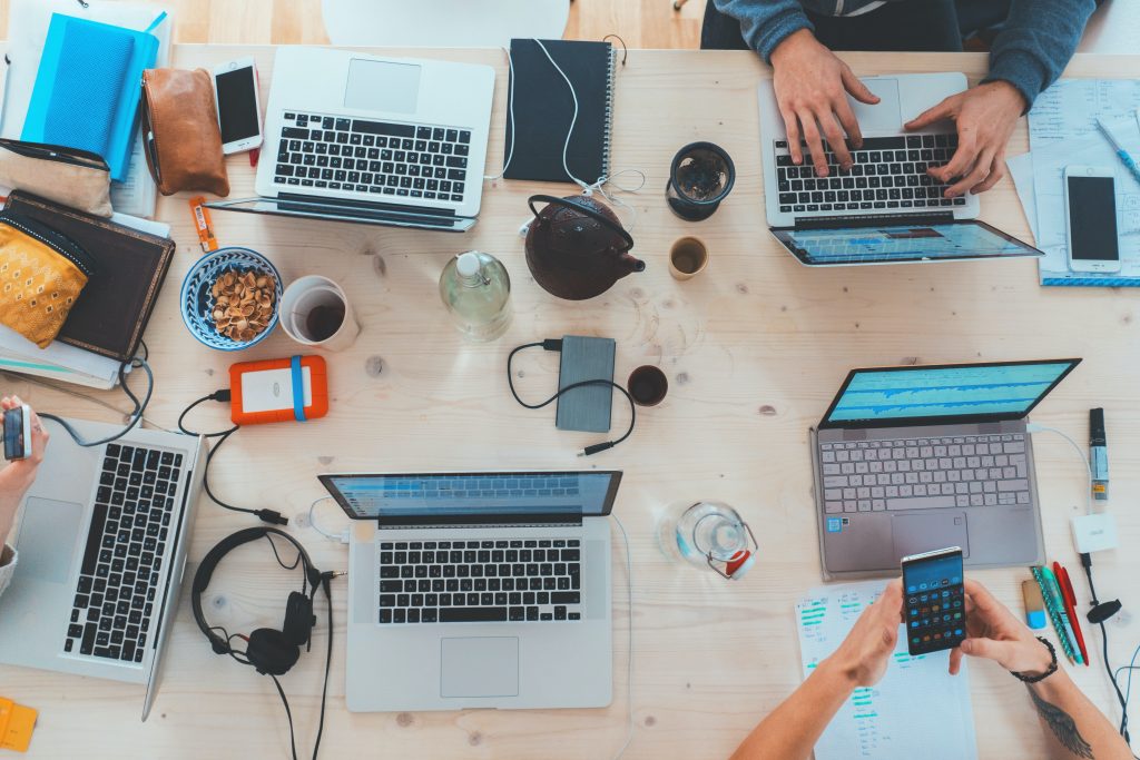 A busy desk full of laptops, phones, drinks and pens etc.