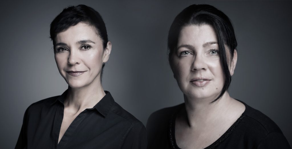 Zwei Frauen mit dunklem Haar, die dunkle Oberteile tragen, posieren für ein professionell aufgenommenes, sanft beleuchtetes Porträt vor grauem Hintergrund – und verkörpern damit die selbstbewusste Ausstrahlung von Zweithaar Experten Dortmund. Beide blicken mit einem leichten Lächeln und neutralem Gesichtsausdruck nach vorne.