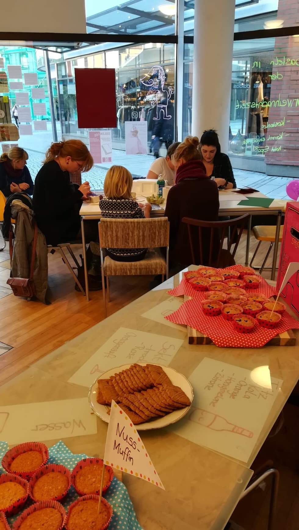 Kinder und Erwachsene sitzen malend an einem Tisch. Im Vordergrund ist Gebäck.